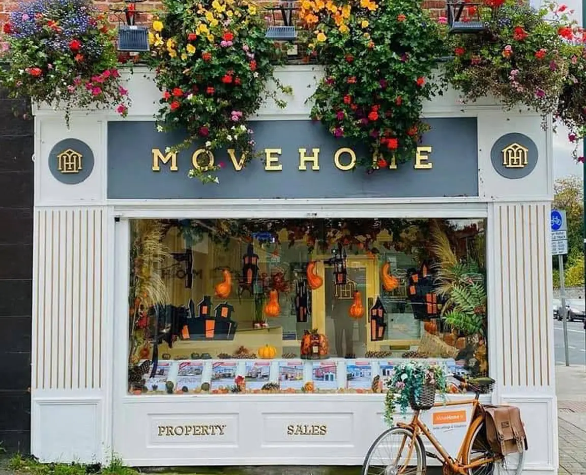 A property shopfront decorated with autumn and Hallowe’en-themed items, pumpkins, and hanging lanterns. Colourful flowers hang above the sign “MOVEHOME.” A bicycle with a basket is parked in front.